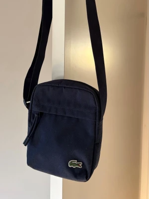  axelväska från Lacoste - Snygg  axelväska från Lacoste i tåligt tygmaterial. Väskan har justerbar axelrem, dragkedja upptill och den klassiska gröna krokodilloggan framtill. Perfekt storlek för det viktigaste och enkel att matcha till olika outfits. Väskan har några färg fläckar som man ser vid sista bilden 