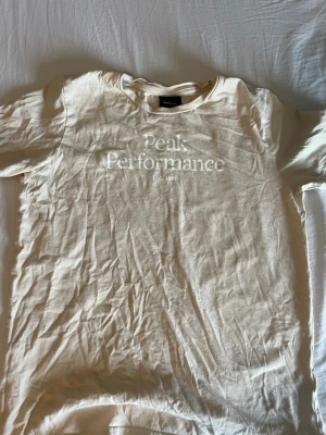 Beige Peak Performance t-shirt - Snygg beige t-shirt från Peak Performance med tryckt logga på bröstet. Klassisk passform och rund halsringning, perfekt till jeans eller shorts. Tillverkad i mjuk bomull för en skön känsla hela dagen.