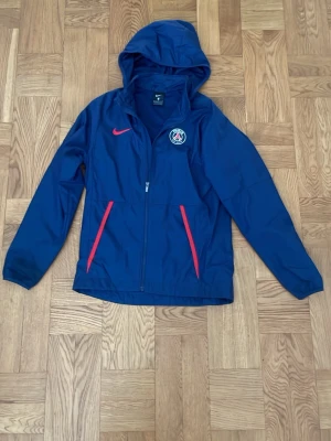 Blå PSG vindjacka från Nike - Snygg blå vindjacka från Nike med PSG-logga på bröstet och röda detaljer vid fickorna. Jackan har huva, dragkedja och meshfoder på insidan. 