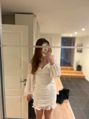 Vit kort omlottklänning med volang - Superfin vit kort klänning med omlottkänsla och volangkant nertill. Klänningen har långa, lätt puffiga ärmar i transparent tyg och en v-ringad halsringning. Sitter figurnära och har snygga rynkade detaljer längs sidorna.