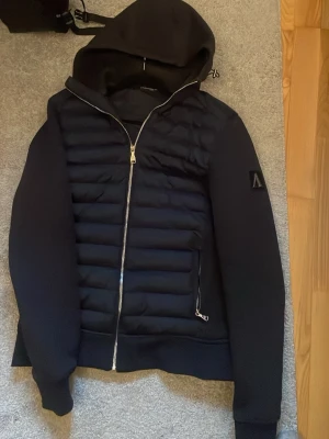 Svart pufferjacka med huva från Moose Knuckles - Snygg svart pufferjacka från Moose Knuckles med quiltad front, dragkedja och huva. Jackan har ribbade muddar, två fickor med dragkedja och en ikonisk logga på ärmen. Perfekt för kalla vinterdagar och riktigt trendig stil.