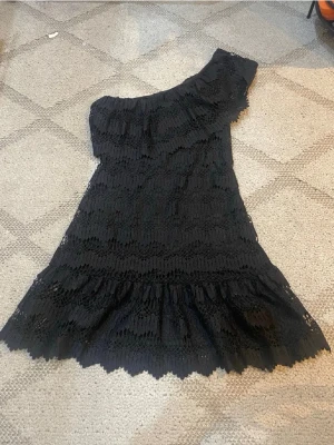 Svart spetsklänning oneshoulder - Snygg svart kortklänning i spets med oneshoulder-design och volangdetalj längst ner. Klänningen har ett feminint snitt och är perfekt för dig som vill sticka ut. Materialet är spets och modellen är figurnära med en trendig asymmetrisk axel. Slutsåld❤️