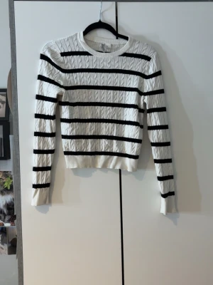 Vit kabelstickad tröja med svarta ränder - Vit kabelstickad tröja från H&M Divided med svarta horisontella ränder. Tröjan har rund halsringning, långa ärmar och ribbade muddar. Perfekt för en stilren och trendig look.