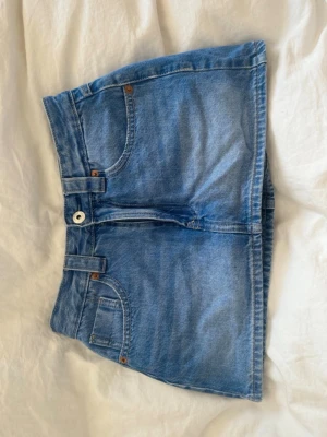 Blå jeanskjol - Snygg blå jeanskjol med klassisk femficksdesign och knappstängning framtill. Kjolen är kort och har en rak passform, perfekt till sommaren. Tillverkad i bomull för en bekväm känsla. Den har dock inbyggda gjorts men gör att klippa bort om man inte vill ha dom och dom har knappt använts.