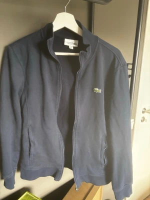 Mörkblå zip hoodie från Lacoste - Snygg mörkblå hoodie från Lacoste med dragkedja framtill och två fickor. Sitter som en Xs-S.  Klassisk krokodillogga på bröstet och ribbade muddar vid ärmslut och nederkant. Tillverkad i mjukt bomullsmaterial för en bekväm känsla. Perfekt för en avslappnad stil.