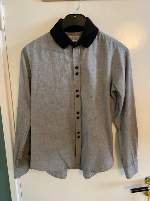 Grå skjorta med svart sammetskrage - Stilren grå bomullsskjorta med svart sammetskrage och svarta klädda sammetsknappar. Skjortan har lång ärm och klassisk passform. Materialet känns mjukt som flanell. Märket (In God We Trust) var ett litet indie-NYC-märke som tyvärr lade ner 2021. 
