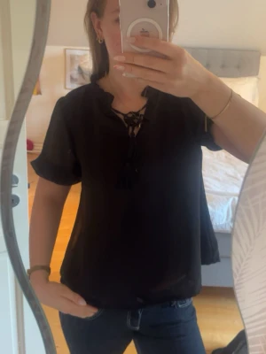 Svart blus med knytning i halsen - Svart blus med korta ärmar och volangkant vid ärmslut. Blusen har en snygg knytning med tofsar vid halsen och är tillverkad i ett lätt, något transparent tyg som ger en luftig känsla. Perfekt att styla med jeans eller kjol.