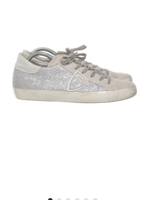 Philippe Model silvriga glitter sneakers - Snygga sneakers från Philippe Model i silverglittrigt material med grå mockadetaljer. Klassisk låg modell med logotyp på sidan och vit sula. Perfekta för dig som vill sticka ut med lite extra bling i vardagen.