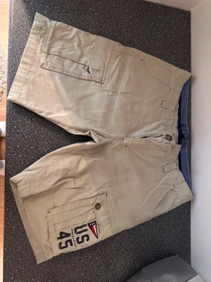 Beige cargo shorts US 45 - Snygga beige cargo shorts från US 45 med flera stora fickor och coolt tryck på ena benet. Perfekta för sommaren med avslappnad passform och praktiska detaljer. Klassisk design med knapp och dragkedja framtill.