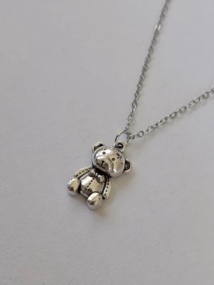 Halsband med nallebjörn i silverfärg - Sött tunt halsband (ankarlänk) med en detaljrik nallebjörn som hänge. Kedjan och hänget är i silverfärgad metall och nallen har en liten rosett runt halsen. Perfekt accessoar för dig som gillar gulliga och lekfulla smycken. Materialet rostfritt stål. 