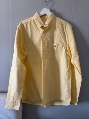 Gul bomullsskjorta från Gant helt ny  - Klassisk gul skjorta från Gant i bomull med lång ärm och bröstficka med broderad logga. Skjortan har knappar framtill och en avslappnad passform, perfekt för en stilren look.