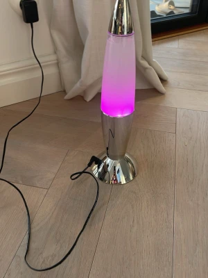 Lavalampa med silverfot och LED - Säljer en stilren lavalampa med silverfärgad metallfot och topp. Lampan har en vit kropp som lyser i lila när den är påslagen. Drivs med sladd och adapter som medföljer. Perfekt som cool inredningsdetalj till rummet.