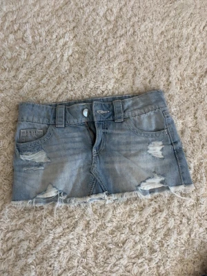 Ljusblå jeanskjol med slitningar - Trendig ljusblå jeanskjol med slitna detaljer och fransig nederkant. Kjolen har klassiska fickor fram och bak samt bälteshällor. Perfekt för dig som gillar en avslappnad och cool stil. aldrig använd, men har ej prislapp kvar så därför kan jag ej trycka på nytt skick. men pga aldrig använd så ser den helt ny ut som att du köpt den i butik!