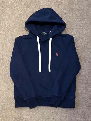 Mörkblå hoodie från Polo Ralph Lauren - Mörkblå hoodie med vit dragsko och klassisk röd Polo-logga broderad på bröstet. Mjuk bomullskänsla, känguruficka framtill och ribbade muddar vid ärmslut och nederkant. Perfekt för en chill och stilren look.