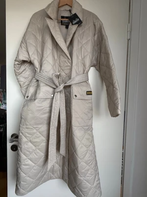 Beige quiltad kappa från Barbour - Stilren beige quiltad kappa från Barbour med bälte i midjan och stora slag. Kappan har tryckknappar vid fickorna och ett klassiskt Barbour-märke på ena sidan. Perfekt för kyliga dagar och ger en trendig look.