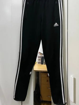 Svarta Adidas träningsbyxor - Svarta träningsbyxor från Adidas med klassiska vita ränder längs sidorna och dragkedjor vid fickorna. Byxorna har en smal passform och resår i midjan. Perfekta för träning eller chill. Adidas-logga på låret.