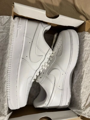 Nike Air Force 1 vita sneakers - Klassiska Nike Air Force 1 sneakers i helvitt läder med perforerad tå, vit sula och vita skosnören. Ikonisk Swoosh-logga på sidorna och metallbricka med 'AF1' på snörningen. Perfekta för en clean och stilren look.