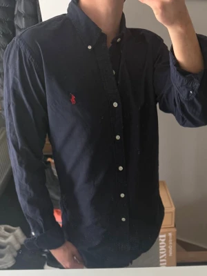 Mörkblå skjorta från Ralph Lauren - Klassisk mörkblå skjorta från Ralph Lauren med vit knappstängning och röd broderad logga på bröstet. Skjortan har button-down krage och långa ärmar. Perfekt för en stilren look. Den har blivit lite missfärgad i armhålorna, därav priset