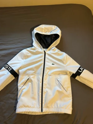 Vit sommar/vår jacka från Lacoste - Snygg vit sommar/vår jacka från Lacoste med svart dragkedja och svarta detaljer på ärmarna där Lacoste-loggan står. Jackan har huva, två snedställda fickor och elastiska muddar. Strlk S/XS. Helt nyskick