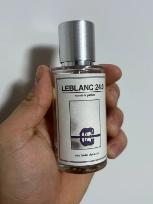 LEBLANC 24.0 parfym - Parfymflaska från one bold chemist med namnet LEBLANC 24.0. Flaskan är cylindrisk i genomskinligt glas med en stilren, silverfärgad metallkork. Etiketten är vit med svart text och en grafisk illustration i lila. Modern och minimalistisk design.