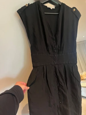Sandro klänning  - Stilren kort svart linne klänning med knappar framtill och markerad midja. Klänningen har korta ärmar, v-ringning och två fickor framtill. Passar perfekt för dig som gillar en klassisk look. Sval klänning för sommaren