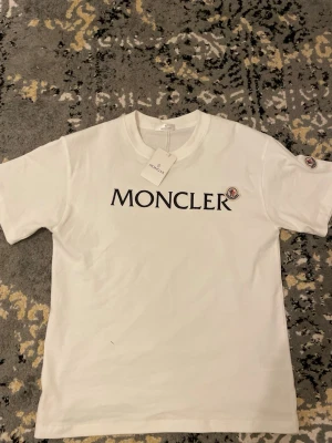 Vit Moncler t-shirt med logga - Snygg vit t-shirt från Moncler med klassisk svart logotyp på bröstet. T-shirten har rund halsringning och korta ärmar. Perfekt för en stilren och trendig look.