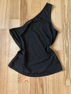 Svart oneshoulder topp  - Aldrig använt,Snygg svart oneshoulder topp i stretchigt material. Toppen har en asymmetrisk axel och ärmlös design som ger en trendig look. Perfekt att styla med jeans eller kjol för en cool outfit.