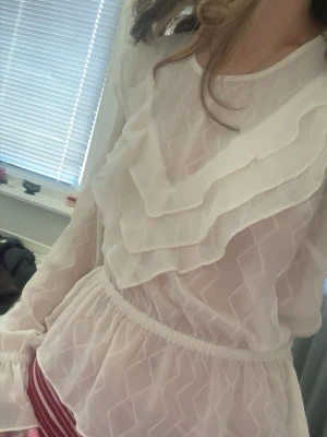 Vit volangblus från Costes - Säljer en superfin vit blus från Costes med transparent material och volangdetaljer framtill. Blusen har lång ärm, resår i midjan och ett lätt mönster i tyget. Perfekt för dig som gillar romantisk och trendig stil. Passar s