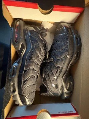 Nike Air Max Plus svarta sneakers - Nike Air Max Plus sneakers i helsvart med mesh och syntetiska detaljer. Snygg vågig design på ovandelen, synlig Air Max-dämpning i sulan och Tn-logo under sulan. Perfekta för dig som gillar streetwear och sportig stil.