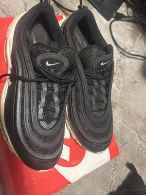 Nike Air Max 97 svarta sneakers - Nike Air Max 97 sneakers i svart med vita detaljer och synlig Air-sula. Ovandelen är i mesh och syntet med vågformade linjer och små Swoosh-loggor på sidan och plösen. Klassisk snörning och bekväm dämpning i sulan.