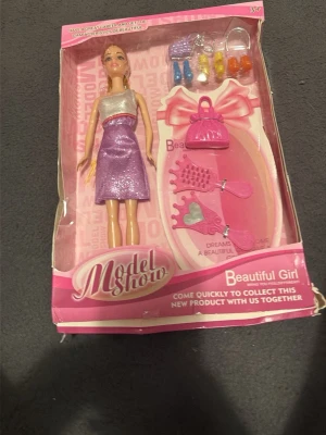 Dockset med lila klänning och tillbehör - Dockset med en docka i glittrig lila klänning och silverfärgat liv. Medföljer rosa handväska, hårborste, tiara, spegel och små accessoarer i olika färger. Perfekt för lek och styling. Allt levereras i originalförpackning.