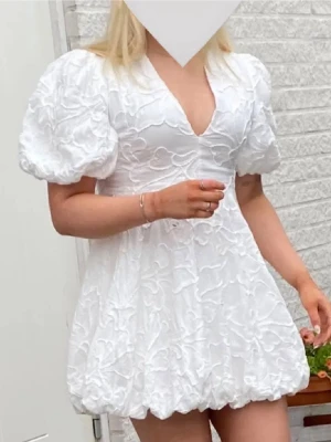 Vit puffärm klänning med struktur - Superfin vit klänning med puffärmar och djup v-ringning. Klänningen har en kort, voluminös kjol med bubblig fåll och ett vackert strukturerat blommönster i tyget. Perfekt för dig som vill sticka ut med en trendig och söt look.