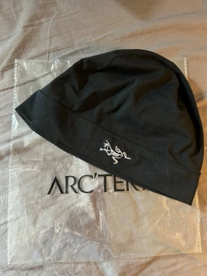 Svart mössa från Arc'teryx - Svart skullcap från Arc'teryx med deras ikoniska logotyp broderad i vitt framtill. Mössan har en enkel och stilren design, tillverkad i ett mjukt och stretchigt material som sitter skönt på huvudet. Perfekt för dig som gillar outdoor och streetstyle.