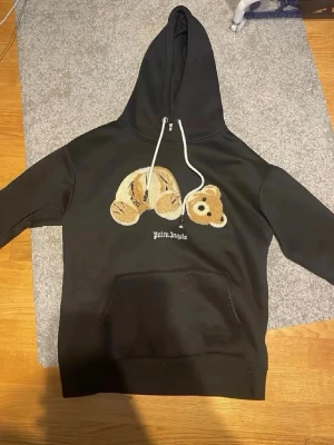 Palm Angels svart hoodie med nalle - Svart hoodie från Palm Angels med stor broderad nallebjörn på bröstet och vit logga under. Hoodien har huva med vita dragsnören och en stor magficka. Materialet är mjukt och skönt, perfekt för en avslappnad streetwear-look.
