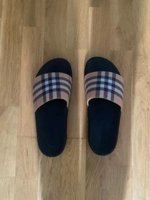 Burberry rutiga slides i svart och beige - Säljer ett par snygga slides från Burberry med klassiskt rutigt mönster i beige, svart, vitt och rött. Tofflorna har svart sula och bred rem över foten i textil med Burberrys ikoniska design. Perfekta för en trendig och avslappnad look.