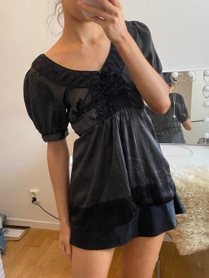 Svart blus från Soaked in Luxury - Snygg svart blus från Soaked in Luxury i glansigt material. Modellen har puffiga korta ärmar, rundad urringning och dekorativa knappar framtill. Blusen är lätt transparent och har en feminin, lös passform.