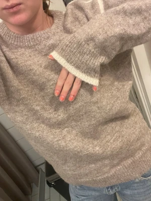 Beige stickad tröja med vit detalj - Mysig stickad tröja i beige med vit rand längs ena ärmen och vid ärmslut. Tröjan har rund halsringning och är långärmad, perfekt för kyliga dagar. Passar till jeans eller kjol för en avslappnad look.