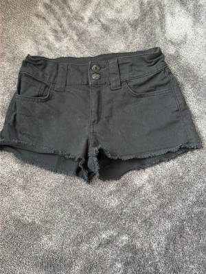 Svarta jeansshorts - Svarta jeansshorts med medel hög midj och råa kanter. Shortsen har två knappar framtill, femficksdesign och bälteshällor. Perfekta för varma dagar och en avslappnad stil. Säljer för att dem inte är min stil längre. De går att göra dem mindre i midjan. 