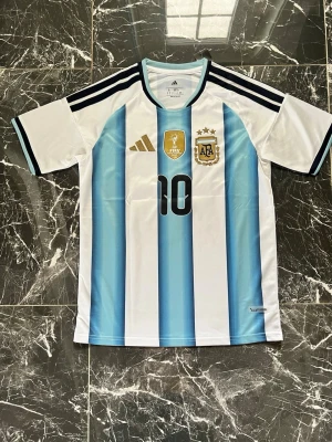 MESSI ARGENTINA VM 2026  - Säljer en officiell Argentina fotbollströja från Adidas med nummer 10. Tröjan har klassiska blå och vita ränder, svarta detaljer och guldiga FIFA 2022-märke samt AFA-emblem på bröstet. Tillverkad i lätt och ventilerande polyester, perfekt för fotbollsträning.