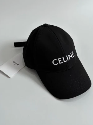 Svart keps från Celine - Stilren svart keps från Celine med vit broderad logotyp framtill. Klassisk böjd skärm och justerbar rem baktill. Tillverkad i ett slitstarkt tyg som ger en snygg och modern look.