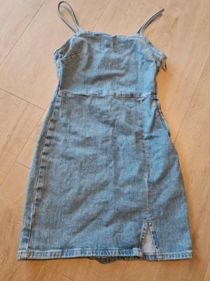 Jeansklänning  - Snygg kort jeansklänning med smala axelband och liten slits framtill. Klänningen har figurnära passform och är tillverkad i klassiskt blått denimtyg. Perfekt för dig som gillar en enkel men trendig look. Använd två gånger bara väldigt fin