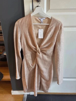 Glittrig champagnefärgad klänning Nelly - Kort glittrig klänning från Nelly  i champagnefärg med lång ärm och v-ringning. Klänningen har en snygg twistdetalj framtill och är figurnära i modellen. Perfekt för dig som vill sticka ut med en elegant look.