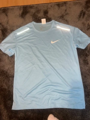 Ljusblå Nike tränings-tshirt - Snygg ljusblå tränings-tshirt från Nike med vitt logotryck på bröstet och reflekterande detaljer på axlarna. Tillverkad i ett lätt och snabbtorkande funktionsmaterial som passar perfekt för träning och sport.