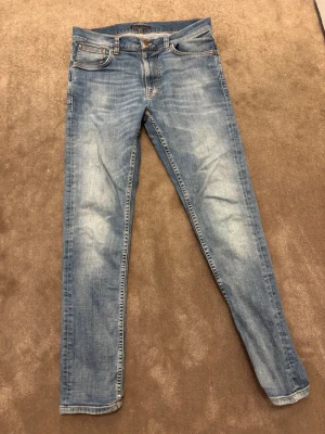 Ljusblå slim fit jeans från nudie - Snygga ljusblå slim fit jeans från nudie med klassisk femficksmodell och slitna detaljer på framsidan. Jeansen har normal midja, dragkedjegylf och är tillverkade i mjuk denim med lite stretch för extra komfort.31/34 storlek
