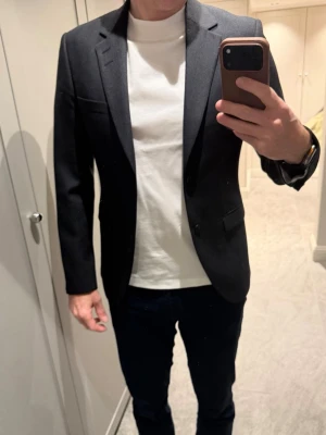 Grå blå kavaj från Tiger of Sweden  - Säljer en stilren grå blå kavaj med slimmad passform och snygga detaljer som knappar på ärmsluten och två framfickor. Kavajen har en bröstficka och är tillverkad i ett material med struktur som ger ett elegant intryck. Perfekt för dig som vill ha en tidlös look. 
