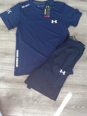 Blått träningsset från Under Armour - Snyggt träningsset med en blå t-shirt och matchande svarta shorts från Under Armour. T-shirten har tryckt logga på bröstet och ärmen samt text längs sidan. Shortsen har elastisk midja och vit logga på benet. Perfekt för gym eller löpning.