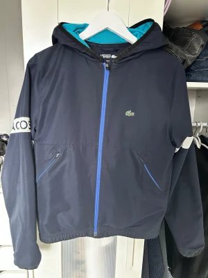 Lacoste windbreaker  - Snygg mörkblå vindjacka från Lacoste Sport med blå dragkedja, huva och turkos meshfoder. Jackan har två fickor med dragkedja och vit detalj med Lacoste-logga på ärmen. Perfekt för blåsiga dagar och sportiga outfits. 16 y 