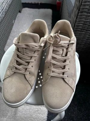 Beige sneakers i mocka-look - Stilrena beige sneakers från VASSU med ovandel i mocka-liknande material och klassisk låg profil. Skorna har matchande beige snörning och vit sula som ger en clean och modern vibe. Perfekta för dig som gillar minimalistisk stil och neutrala färger.
