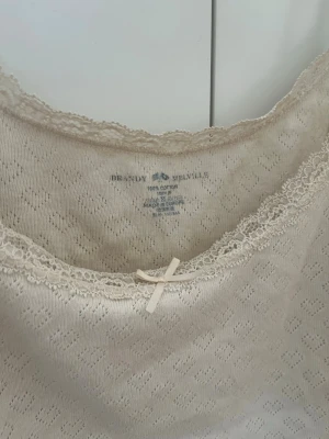 Beige linnetopp från Brandy Melville - Söt beige linnetopp från Brandy Melville med spetskant och liten rosett framtill. Tillverkad i 100% bomull med diskret hålmönster. Perfekt för varma dagar eller som layering-piece.
