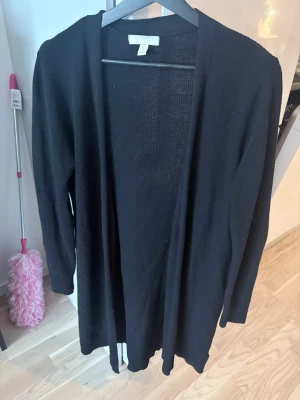 Svart lång kofta från H&M - En svart, lång och öppen kofta från H&M. Koftan har långa ärmar och är tillverkad i ett tunt, mjukt stickat material. Perfekt att slänga över en t-shirt eller topp för en chill och enkel look.